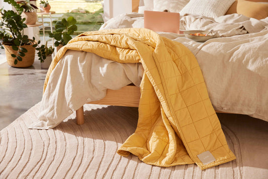 A textured cream rug with timber bed base on top, covered with linen bedding and a yellow throw