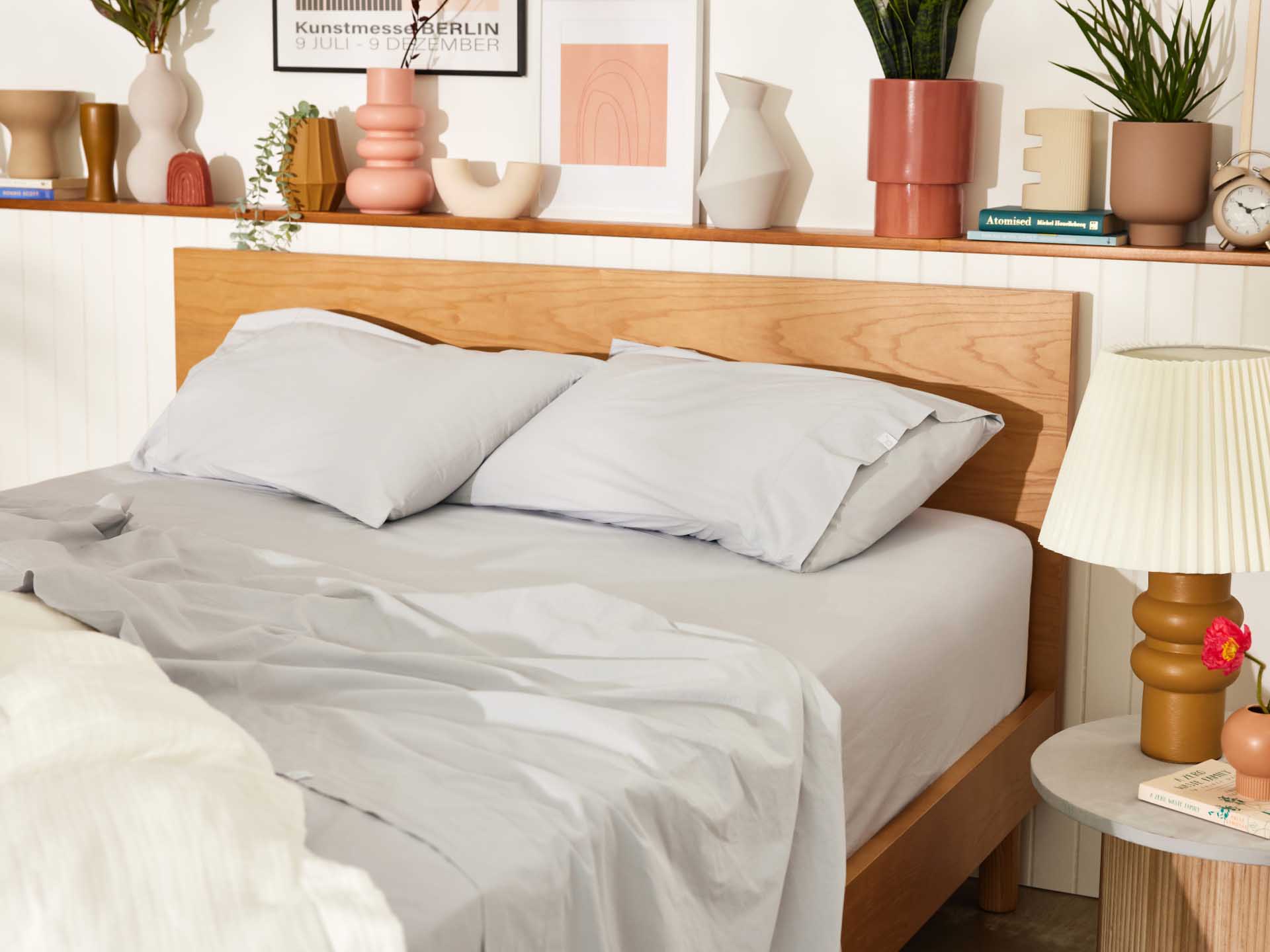 Wooden bed frame with light grey sheets, surrounded by decorative vases, plants, books, and framed art on a shelf. A lamp sits on a side table.
