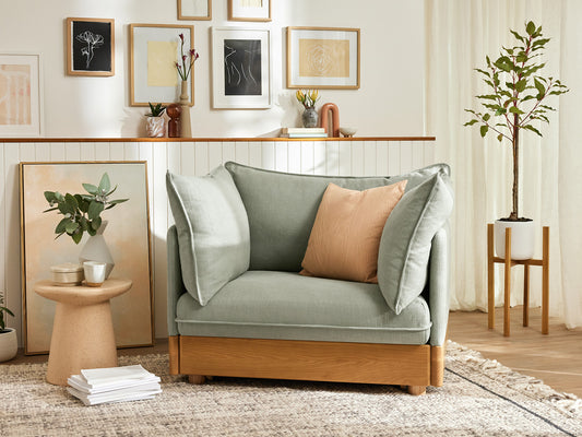 Sage green armchair with a peach cushion, wooden base, next to a small round wooden table. Wall art and plants in a light-filled room.