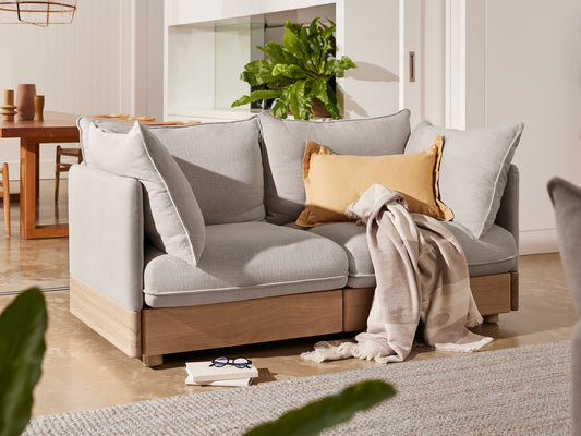 Light grey sectional couch with wooden base, adorned with a mustard cushion and a beige throw. Nearby, a table and plant enhance the cosy setting.