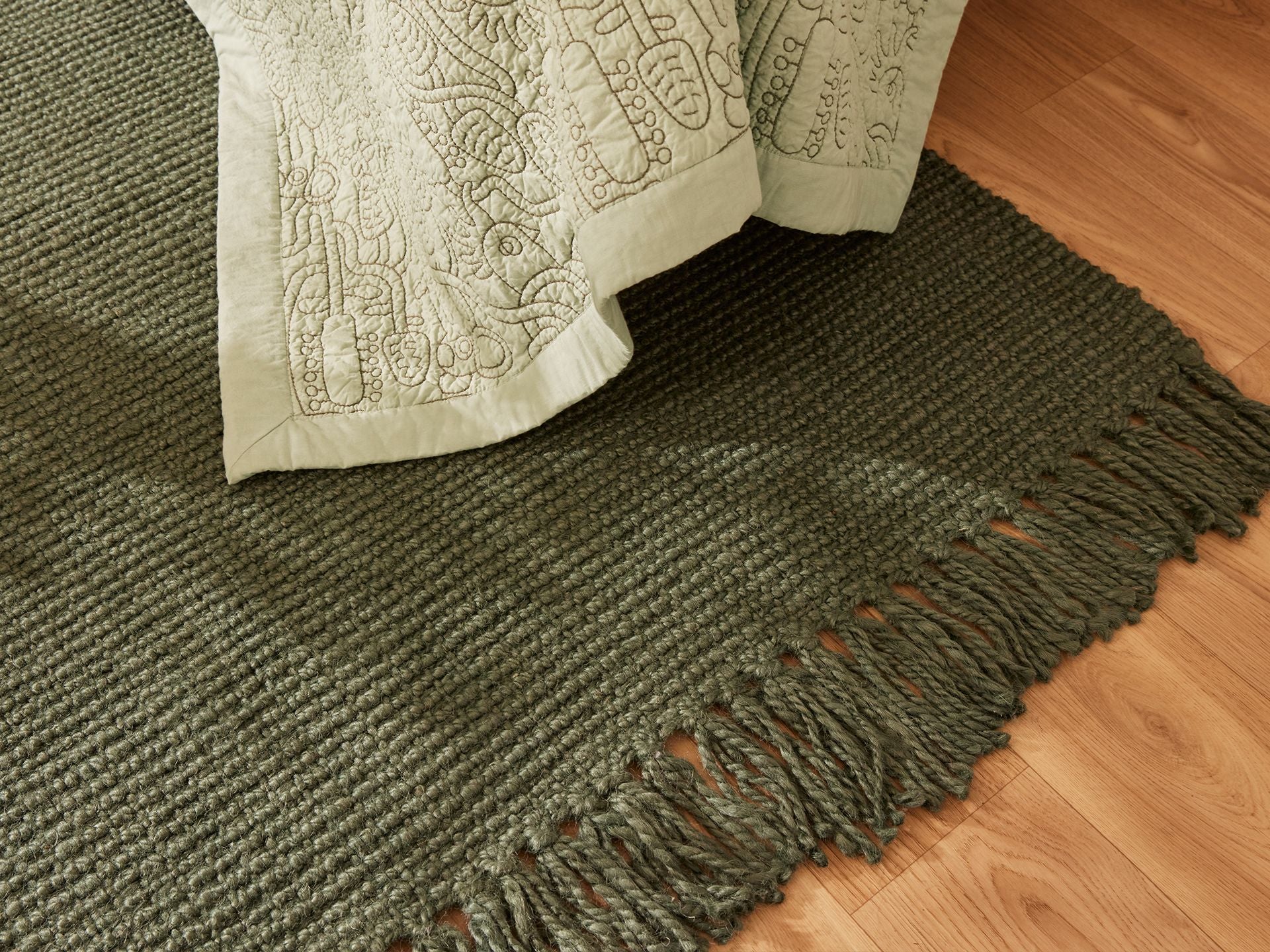 Green woven rug with tassels on a wooden floor, partially covered by a light green embroidered quilt.