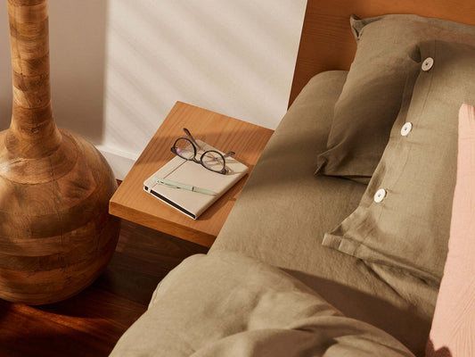 Bed with olive green linen sheets, wooden bedside table with glasses and notebook, and a textured wooden lamp base.