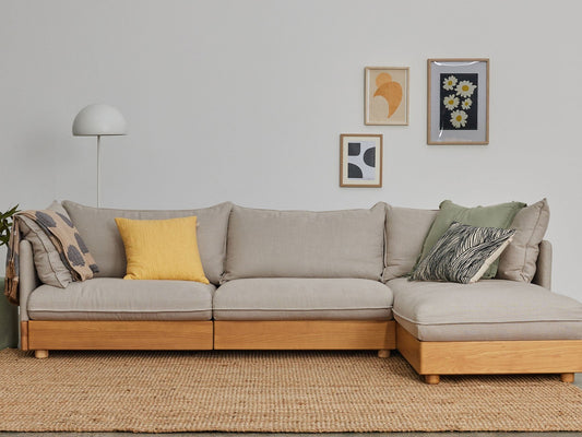 Beige L-shaped sofa with wooden base, colourful cushions, on a woven rug. Minimalist wall art and a white floor lamp in the background.