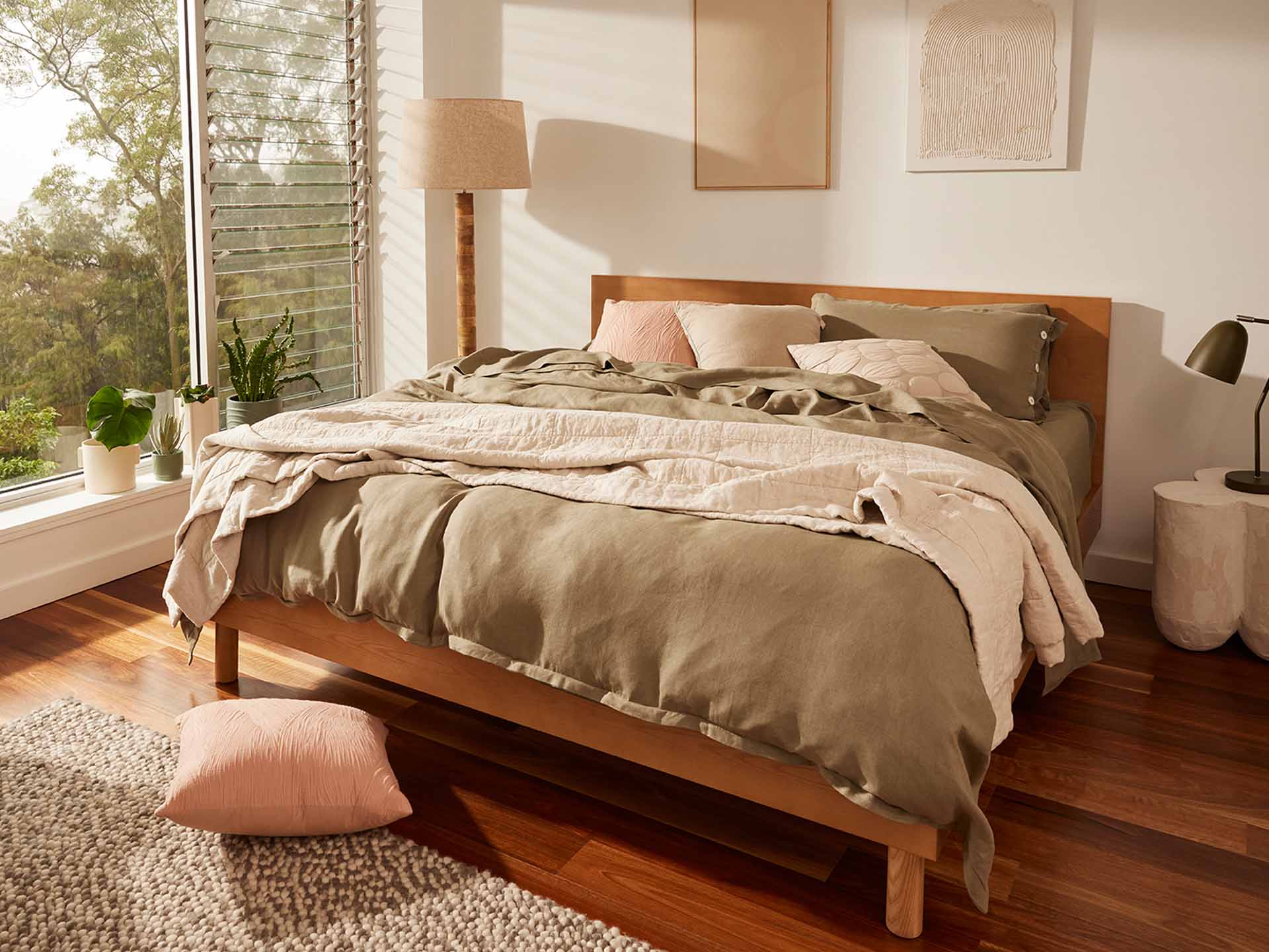 A cosy bedroom with a wooden bed frame, olive green bedding, pink cushions, and a bedside lamp. Sunlight filters through a large window.