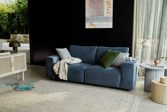 A dark blue sofa with green and grey cushions and a beige throw sits on a light rug. A side table and lamp are nearby; art hangs above.
