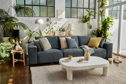 Modern living room with a dark grey sofa adorned with assorted cushions, a white oval coffee table, various plants, and wooden flooring.