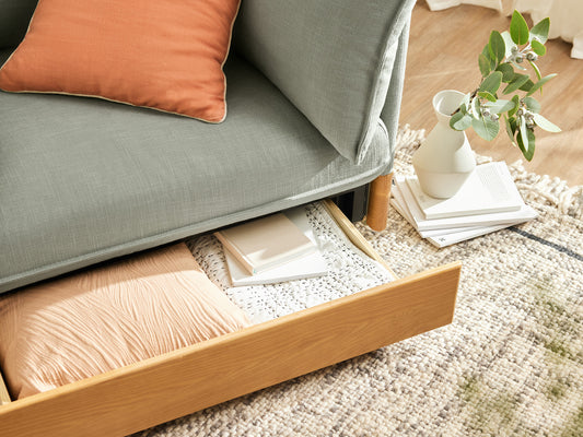 Sage green sofa with peach cushion, open under-seat drawer showing fabric storage. White vase with leaves and books on a woven rug.