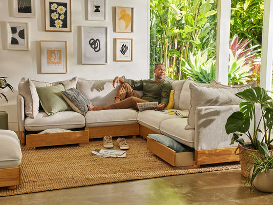Cosy living room with a man on a light grey modular sofa, surrounded by green plants and framed art. Beige rug, and wooden floor.