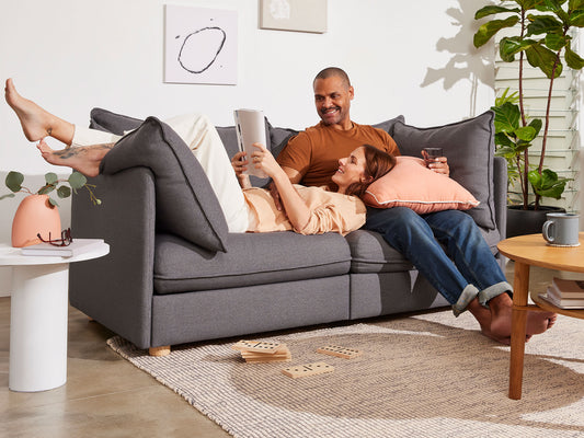 Couple relaxing on a dark grey sofa with plush cushions; a coffee table and plants in the background; soft, neutral decor tones.
