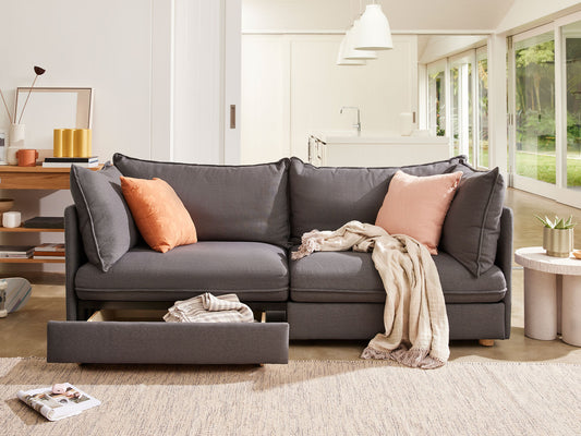 Dark grey sofa with chaise lounge and two orange cushions. Features a pull-out drawer, draped beige throw, and sits on a light beige rug.