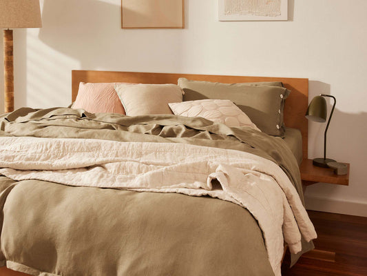 Cosy bedroom with a wooden headboard, olive-green and cream bedding, multiple pillows, and a small green bedside lamp on a wooden nightstand.