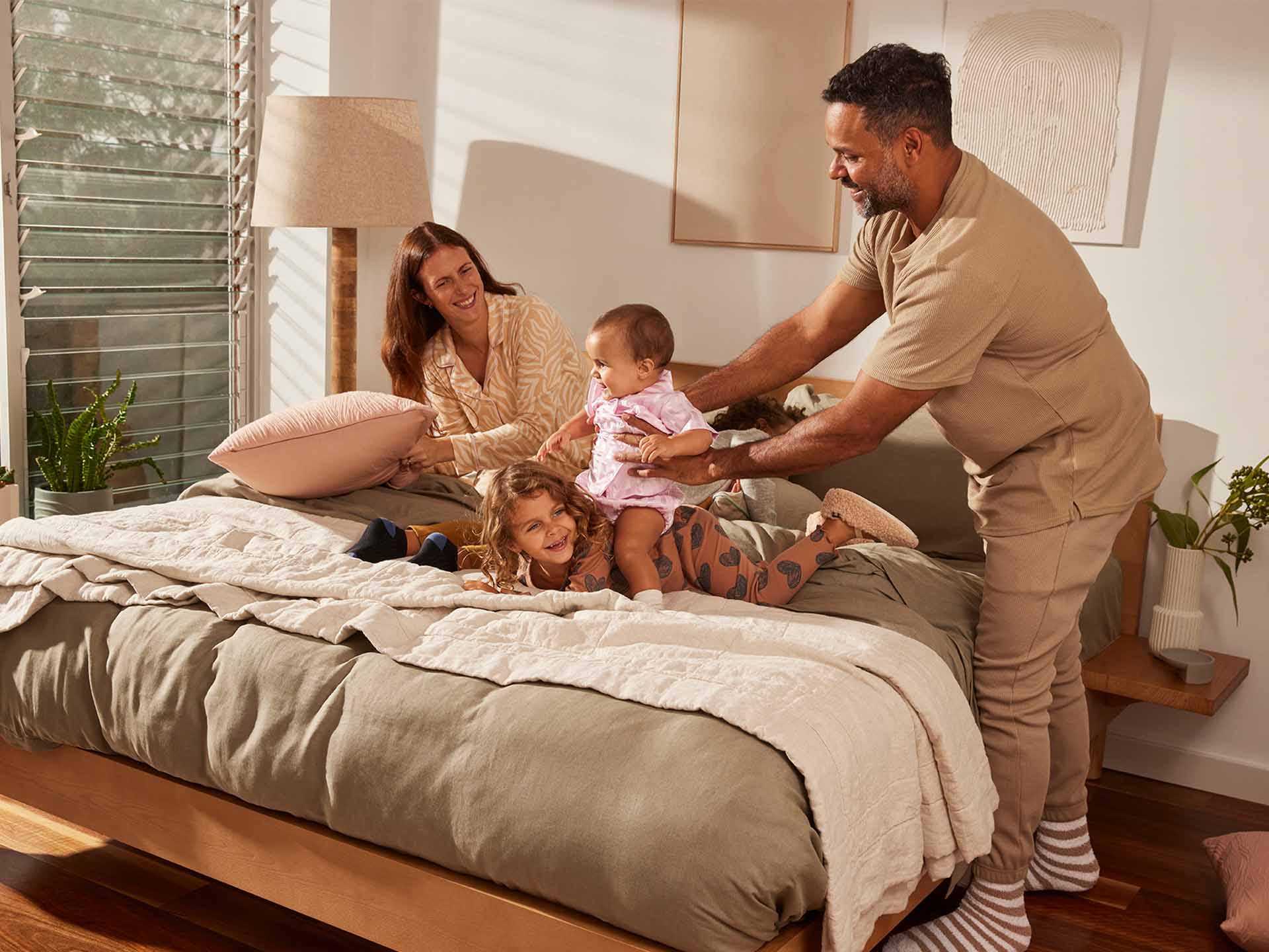 Family playing on a bed with beige bedding, light wood frame, and white cushions. Wooden floor, potted plants, and a lamp in the background.