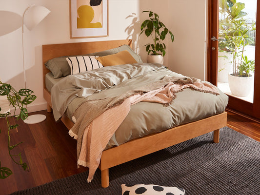 Modern bedroom with a light wood bed, green duvet, and pastel pillows. Features indoor plants, wooden floors, and a grey rug under soft lighting.
