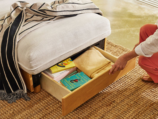 Wooden storage ottoman with white cushion, holding books and a yellow blanket. Striped throw draped over top. Brown woven rug underneath.