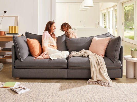 A cosy dark grey sofa with peach cushions and a beige throw. Two people sit comfortably, enjoying a bright, airy living room atmosphere.