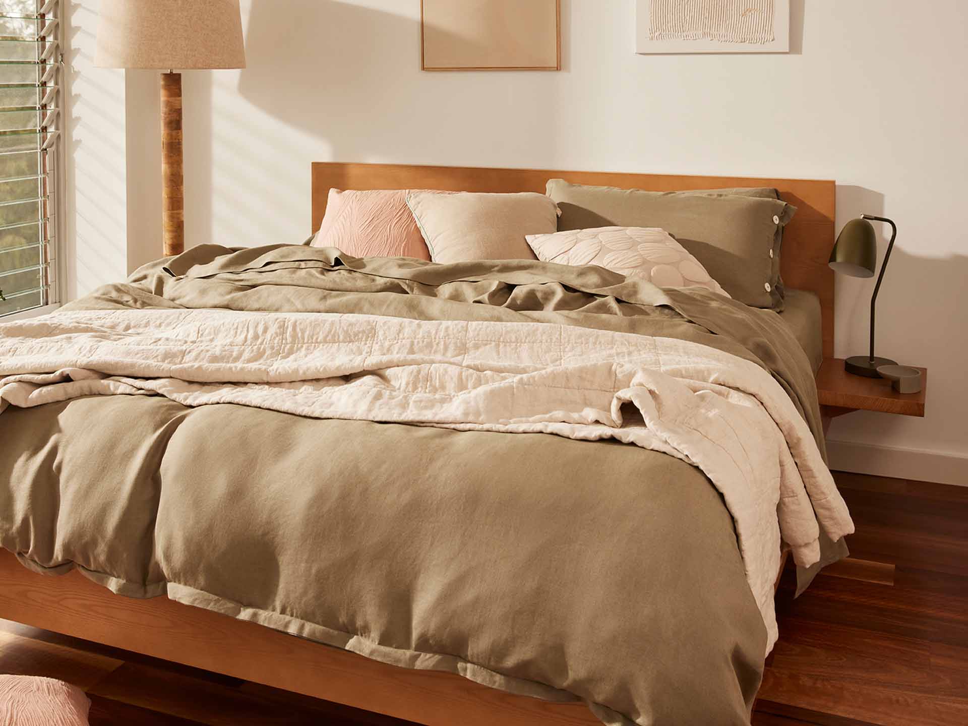 Wooden bed with light brown and cream bedding, pink and grey cushions. Tall lamp and framed art on wall. Wooden floor, natural light.