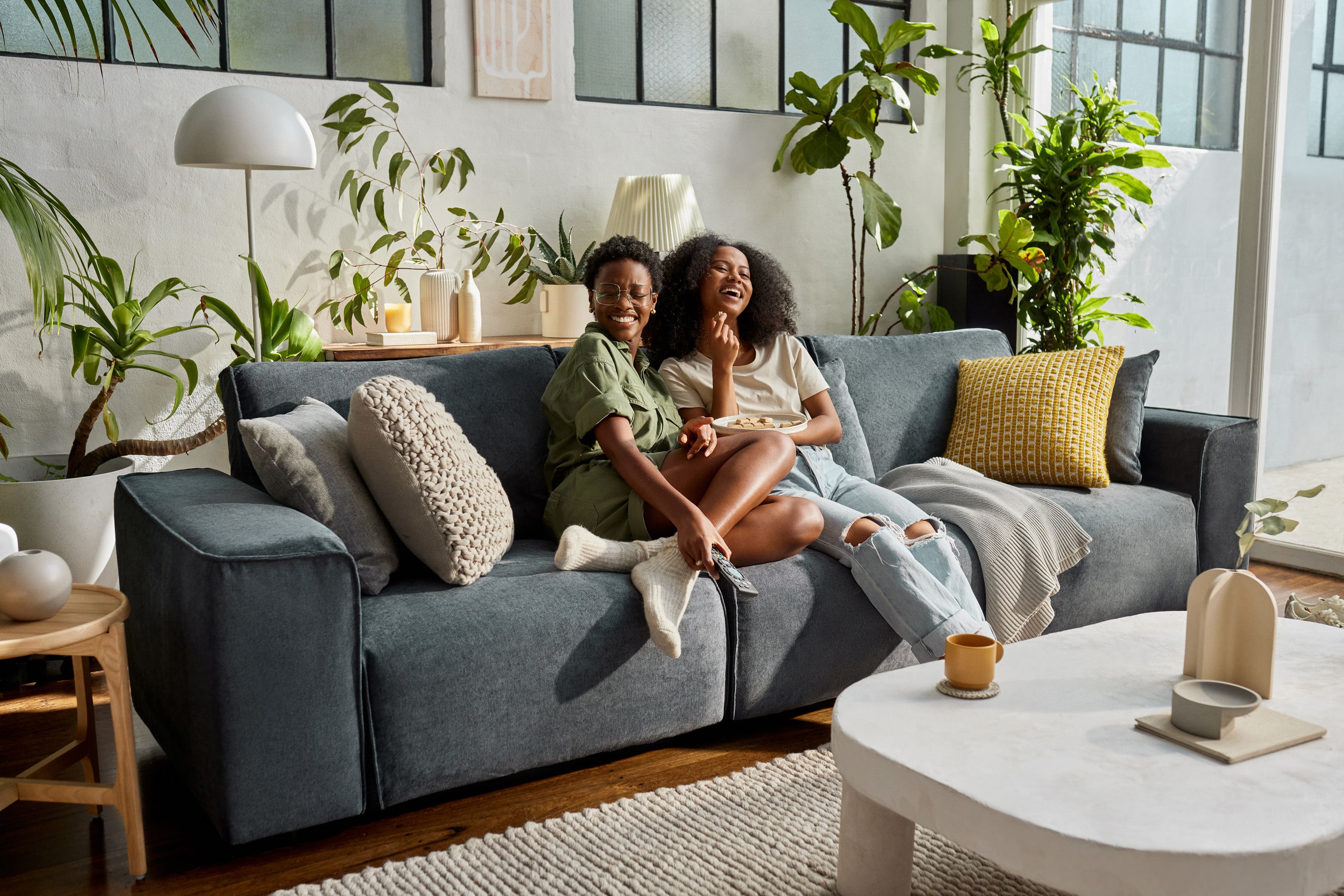 A dark grey sofa with cream and yellow cushions in a living room, surrounded by plants, a white coffee table, and a large window.