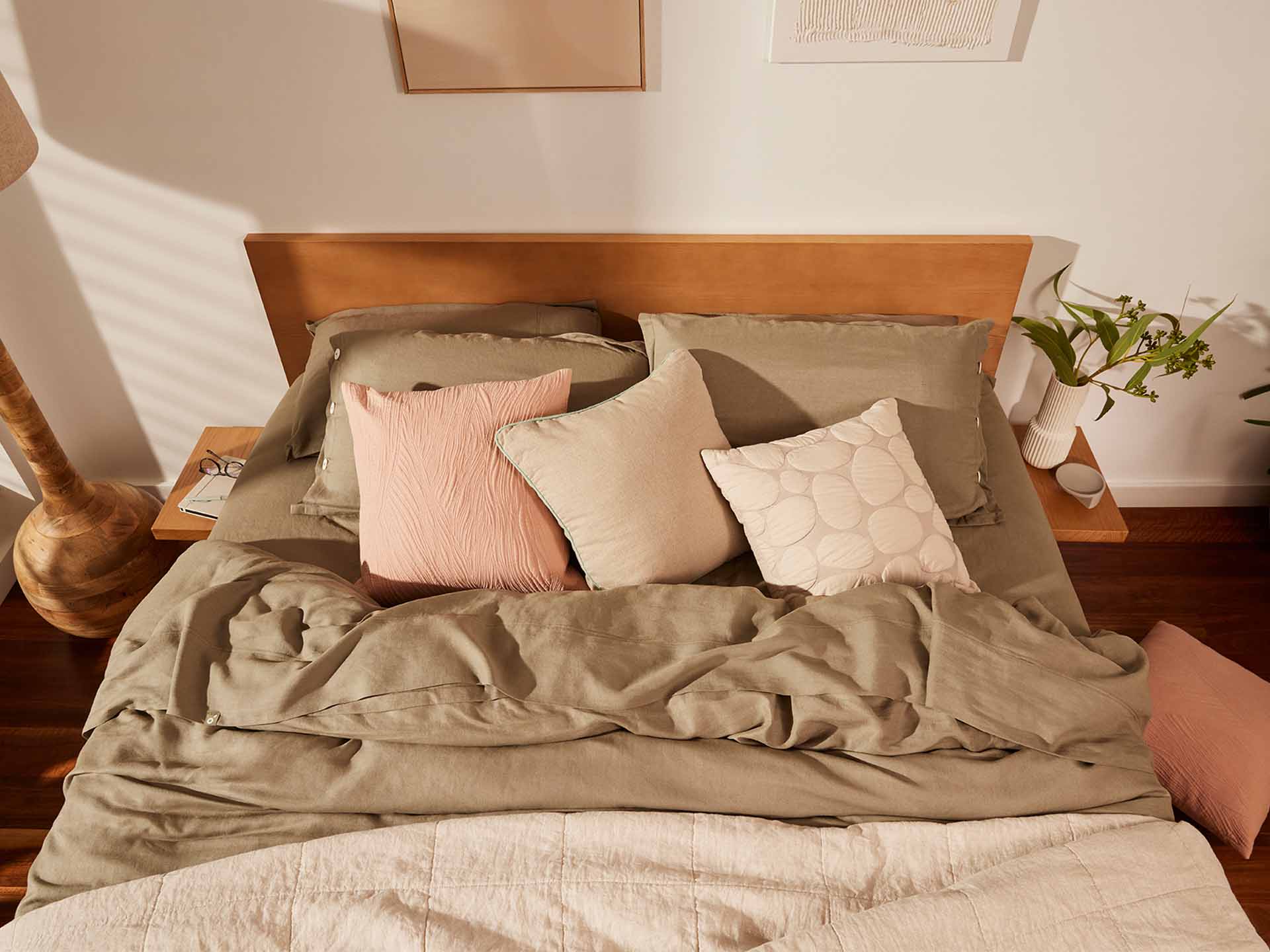 Bed with light brown quilt, beige and pink cushions, wooden headboard, bedside table with plant, and warm lighting on wooden floor.