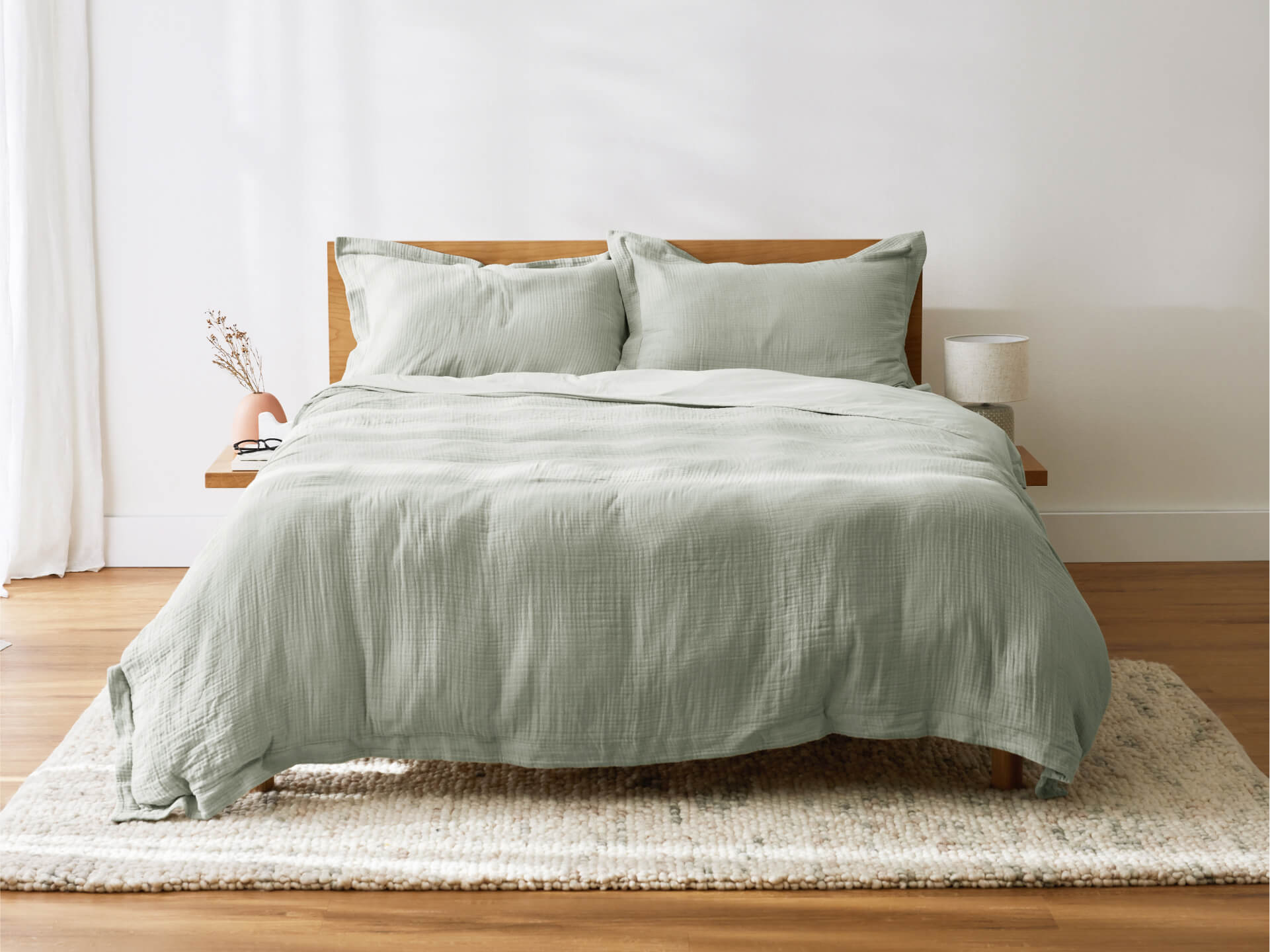 Wooden bed with sage green quilt and pillows on a textured beige rug, in a minimalist room with white walls and sheer curtains.