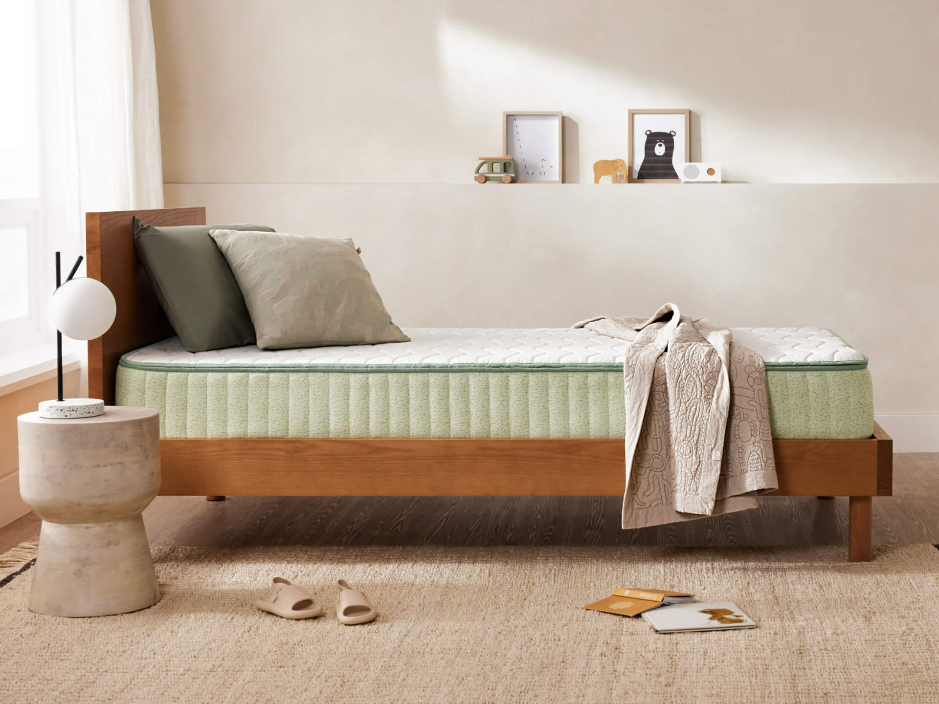 Single bed with light green mattress and pillows, and a beige quilt. Wooden frame, concrete side table, slippers, and minimal décor in a bright room.