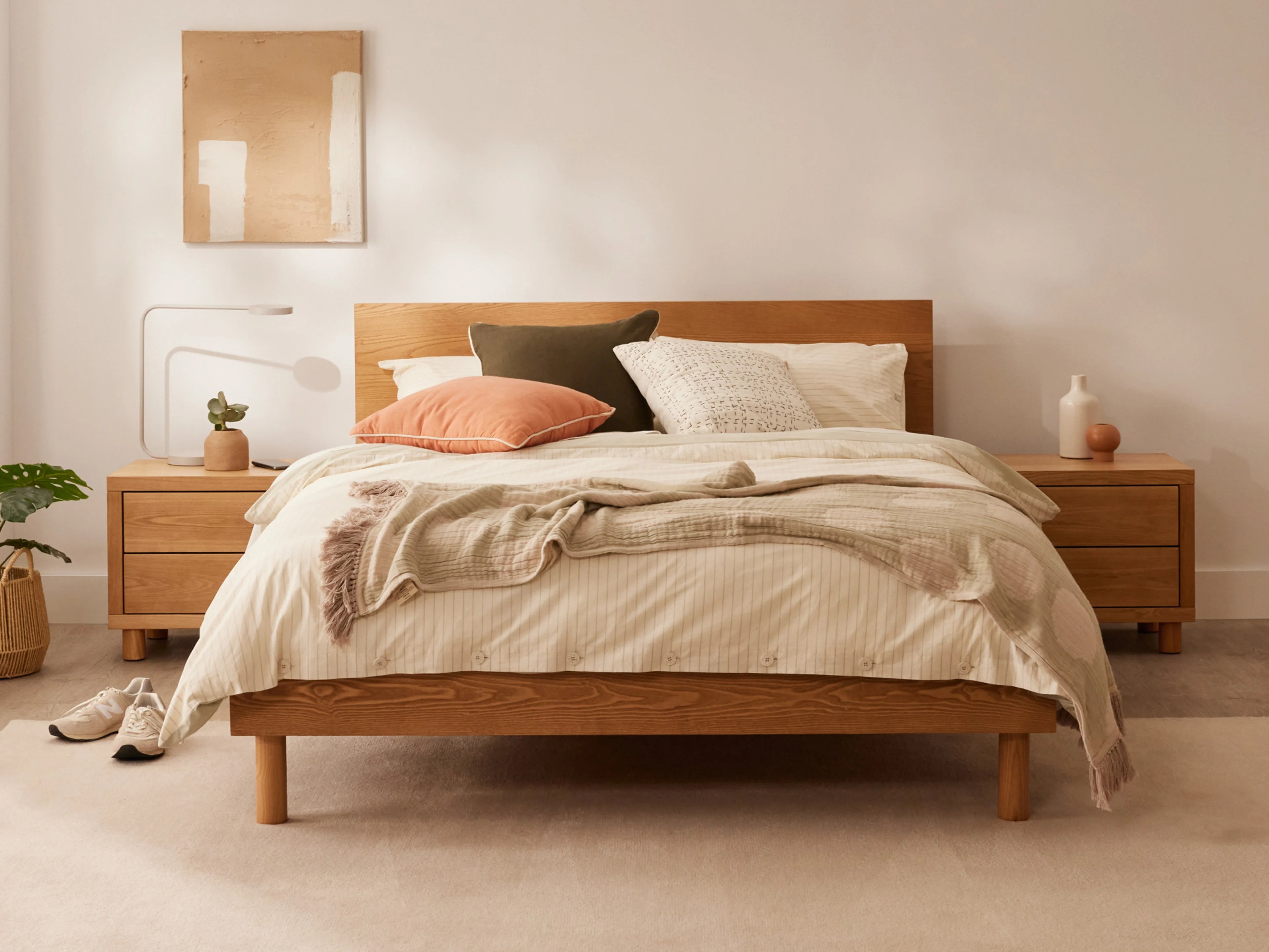 Wooden bed with light beige bedding, cushions in white, brown, and pink. Two wooden bedside tables, wall art, and decor in neutral tones.
