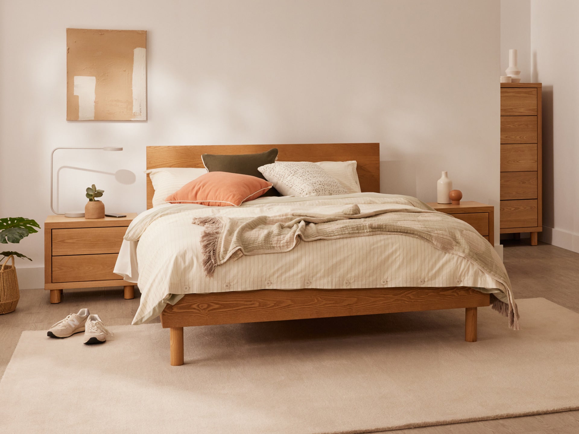 Bedroom with a wooden bed, beige bedding, and cushions. A bedside table with a plant, and drawers. Neutral tones and soft lighting.