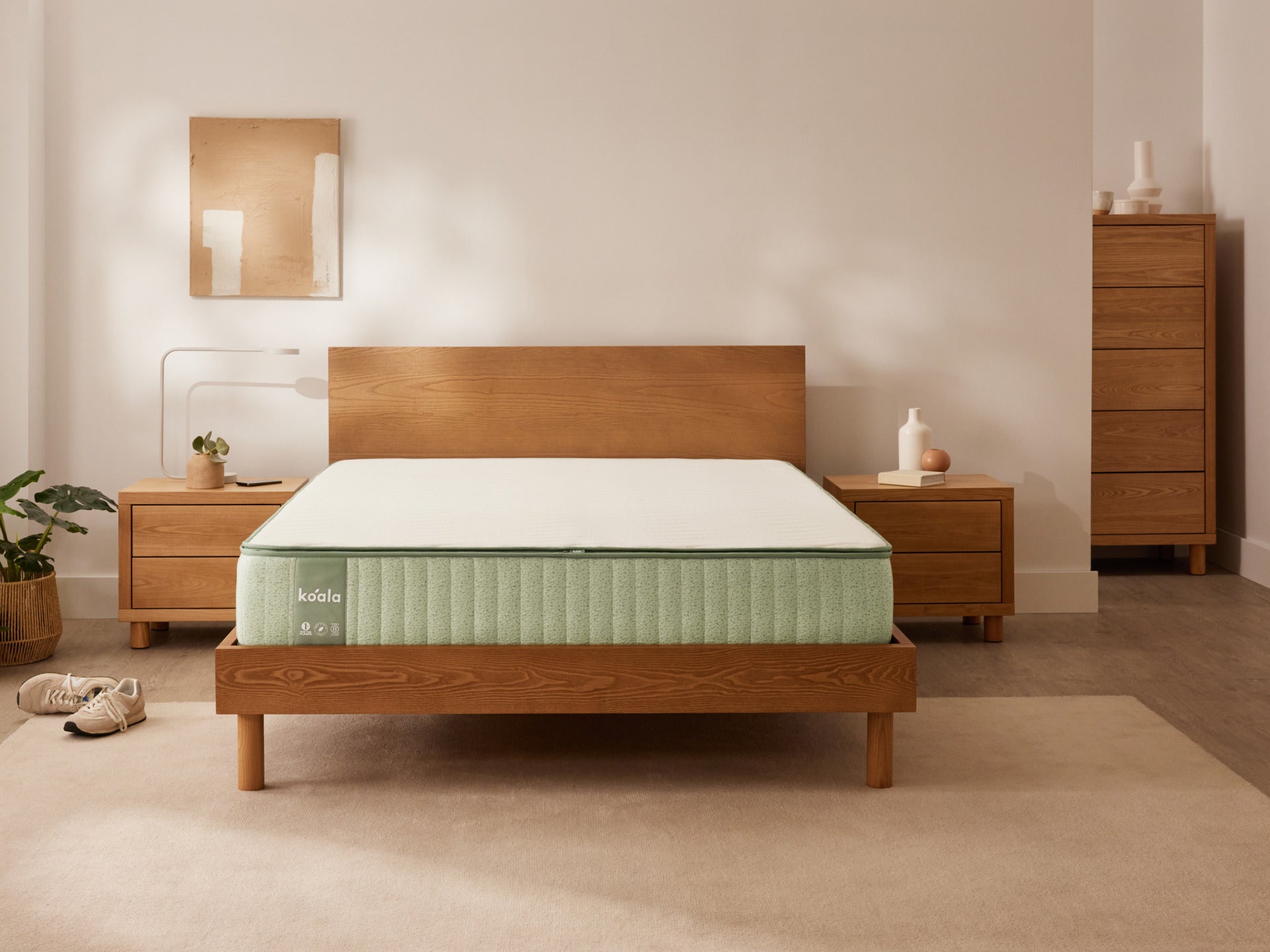 Bedroom with wooden bed frame and light green mattress. Flanked by wooden bedside tables, neutral decor, rug, greenery, and dresser.