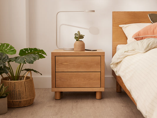 Bedroom with light brown wooden bedside table, potted plant, white lamp, and light-coloured bedding. Green plant in woven basket beside.