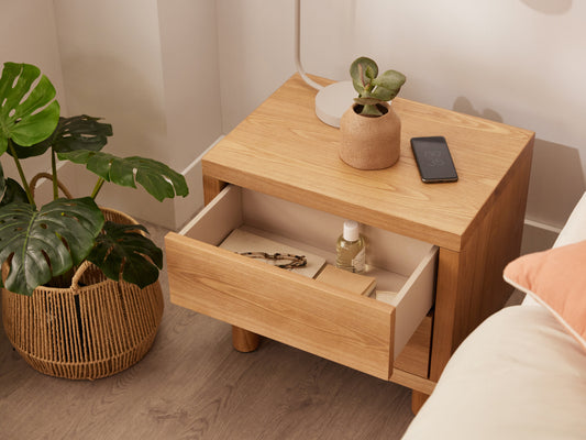 Wooden bedside table with two open drawers, a potted plant, and a smartphone. Next to it, a rattan basket with green leaves on a wooden floor.