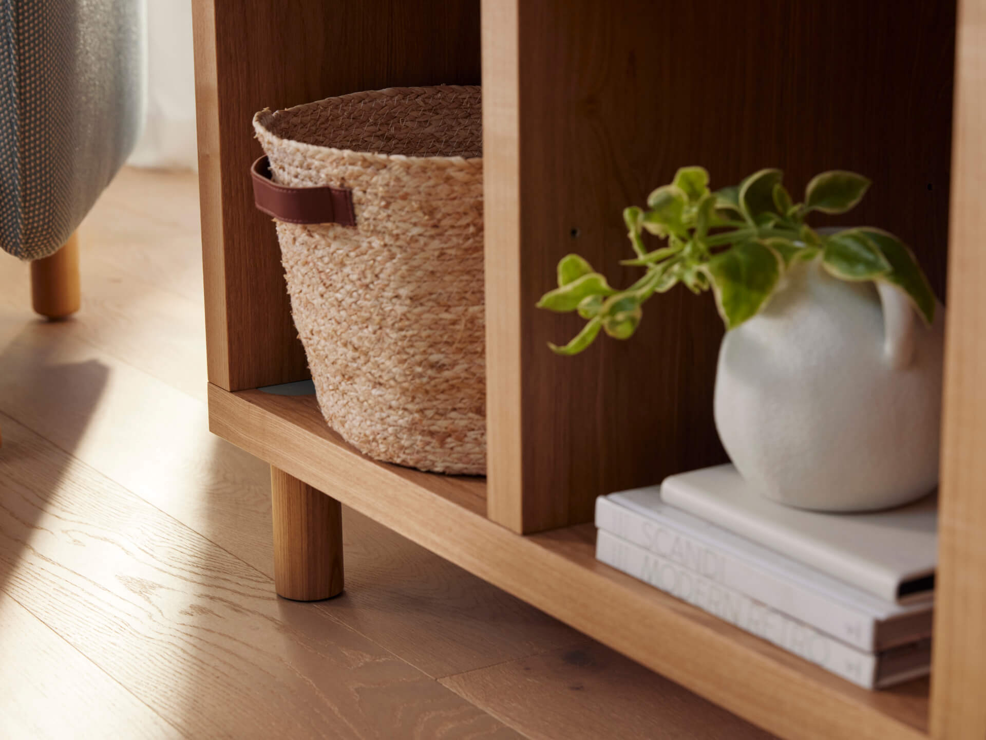 Wooden shelf with woven basket, small potted plant on books. Light wood floor, chair leg partially visible. Cozy interior decor.