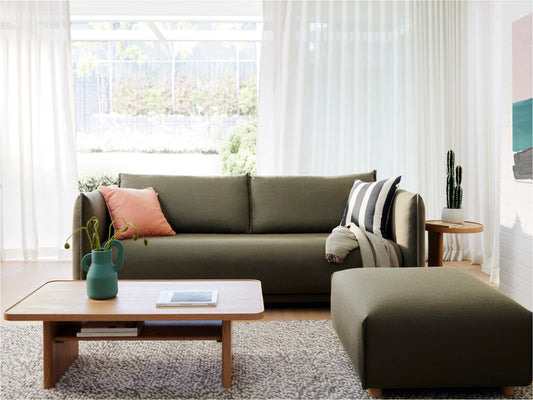 Living room with olive green sofa, matching ottoman, and wooden coffee table on a beige rug. White curtains, plants, and a pink cushion accent.