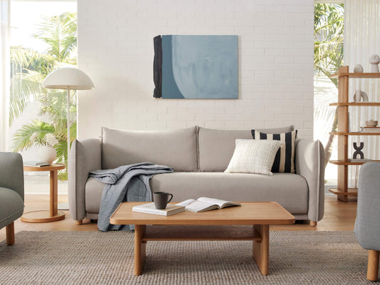Modern living room with a light grey sofa, wooden coffee table, and decor. A floor lamp and abstract painting accent the bright space.