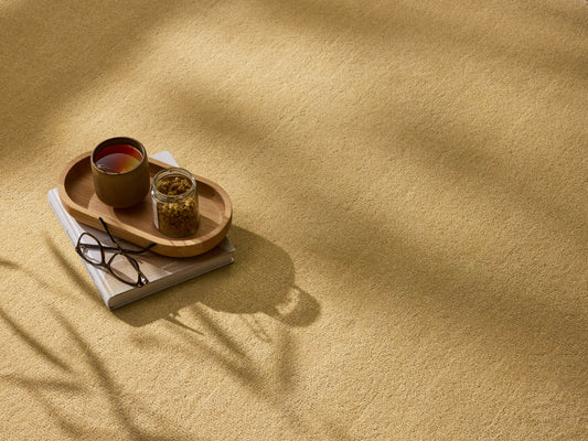 A sandy beige carpet with a wooden tray holding a drink, snack, glasses, and a book. Soft shadows play across the textured surface.