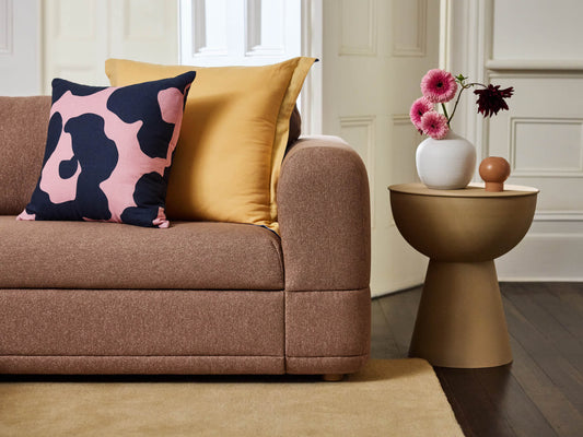 Brown sofa with yellow and pink patterned cushions, next to a round tan side table with pink flowers in a white vase on a wooden floor.