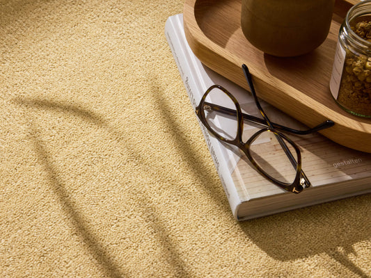 Reading glasses on a closed book beside a wooden tray with a jar on a light brown carpet. Shadows cast a pattern across the scene.
