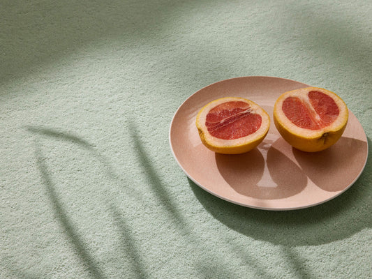 Halved grapefruit on a beige plate, resting on a soft green textured surface with plant shadows.