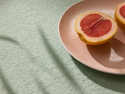 Halved grapefruit on a pink plate, resting on a light green textured surface with shadow patterns.