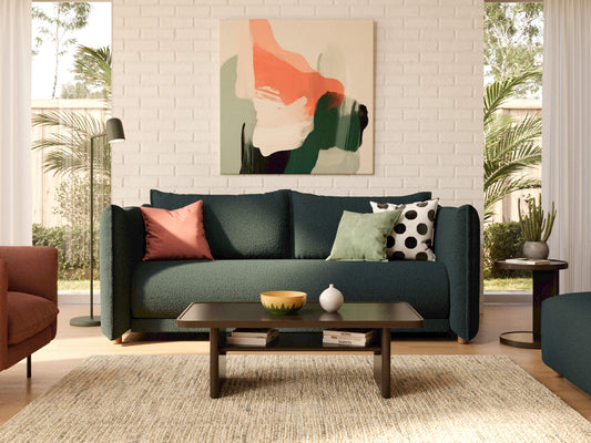 Modern living room with a dark green sofa, colourful cushions, abstract art, and a dark coffee table on a beige rug. White brick wall backdrop.