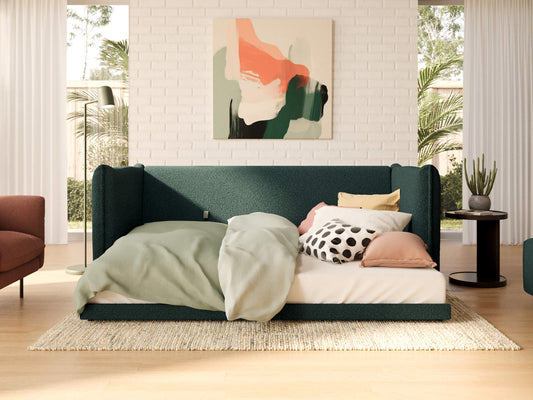 Green upholstered daybed with pillows and blanket, set against a white brick wall. Abstract artwork above; indoor plants and side table nearby.