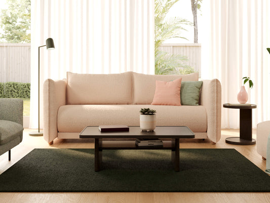 Beige sofa with pink and green cushions, black coffee table on dark rug, floor lamp, and side table with plant against windowed backdrop.