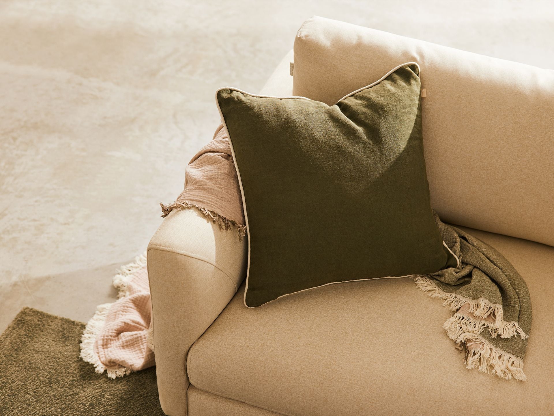 Cream sofa with an olive green cushion and a pink fringed throw draped over the armrest on a light grey textured carpet.