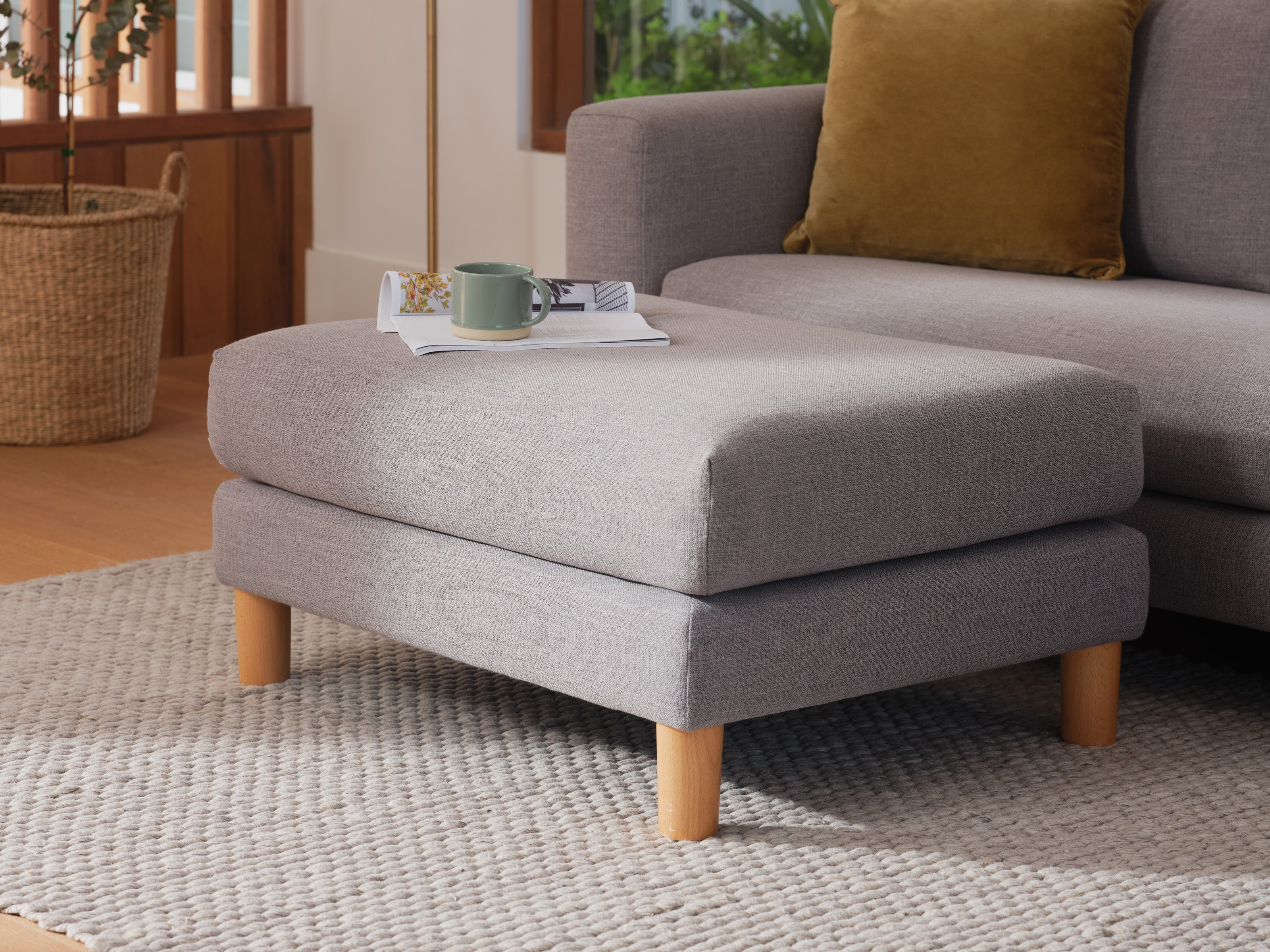 Grey ottoman with wooden legs on light grey rug, near grey sofa with mustard cushion, a cup, and magazines. Wicker basket in the background.