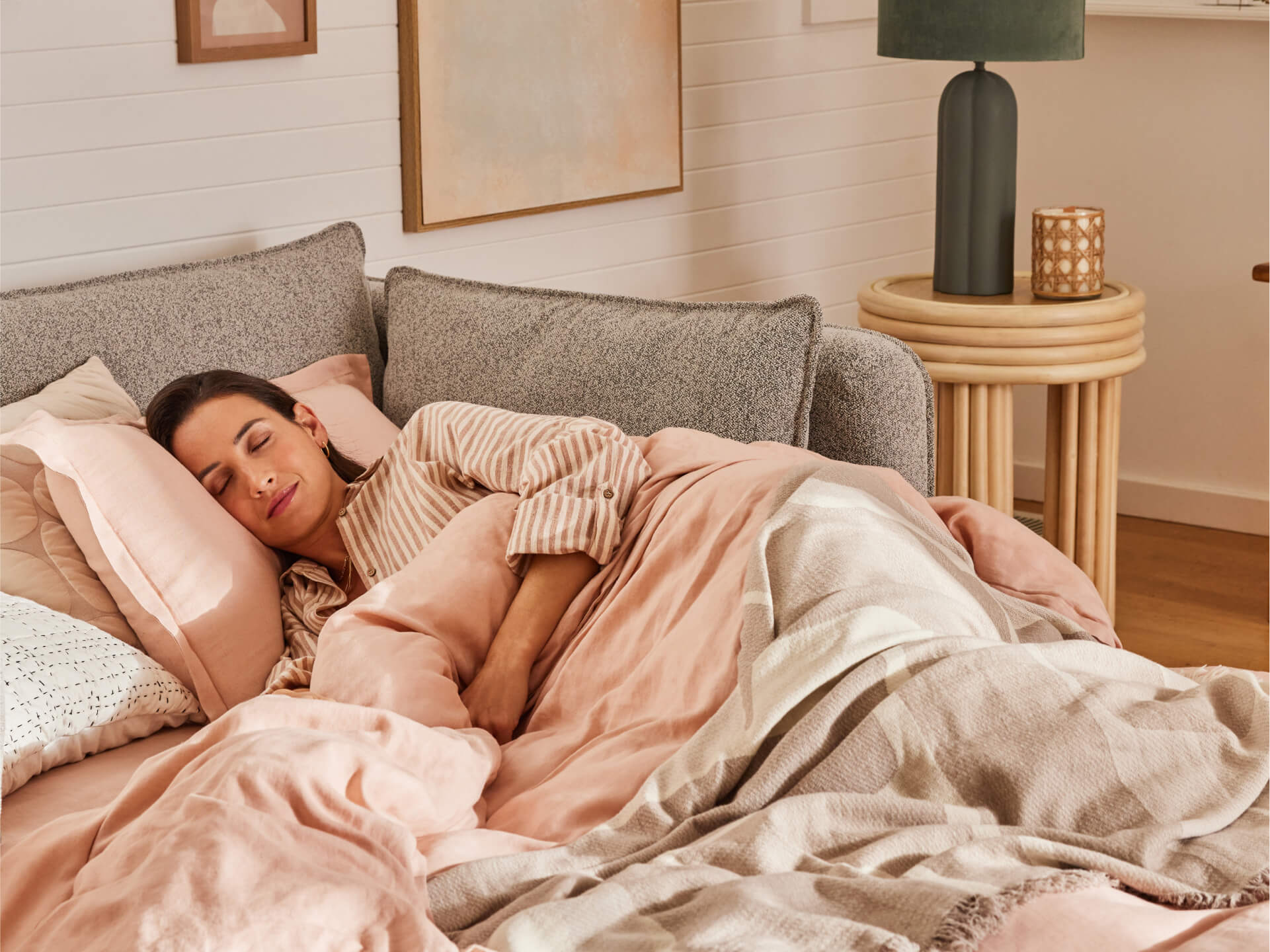 Woman sleeping on a grey couch with pink bedding and striped shirt. Round wooden side table with a green lamp. Cosy and comfortable.