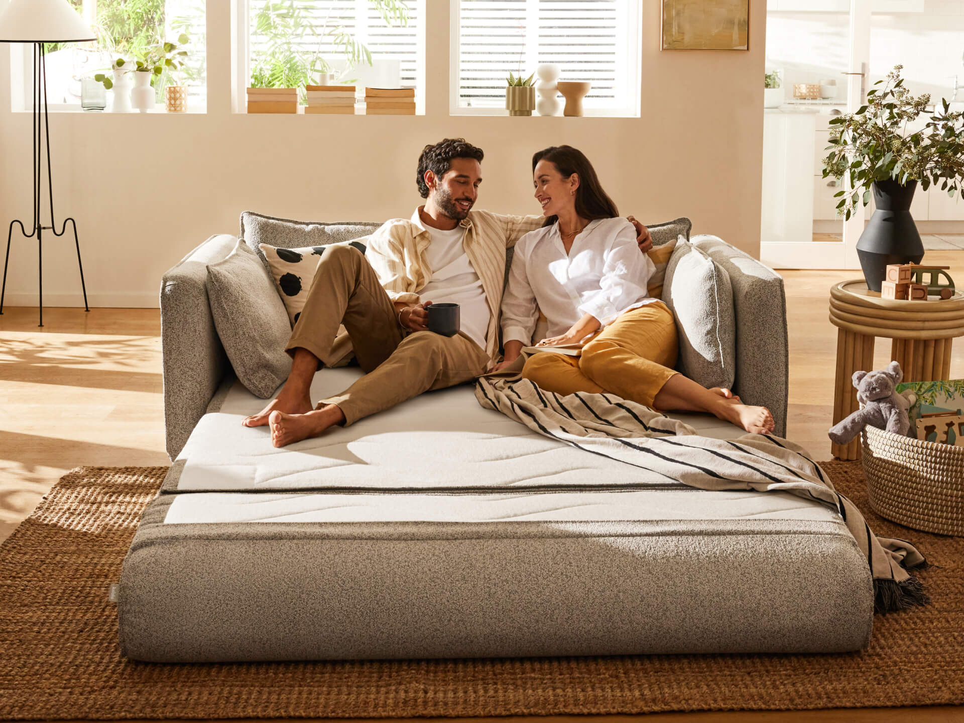 Couple lounging on a light grey sofa bed with cushions, surrounded by natural decor in a sunlit room, featuring a woven rug and indoor plants.