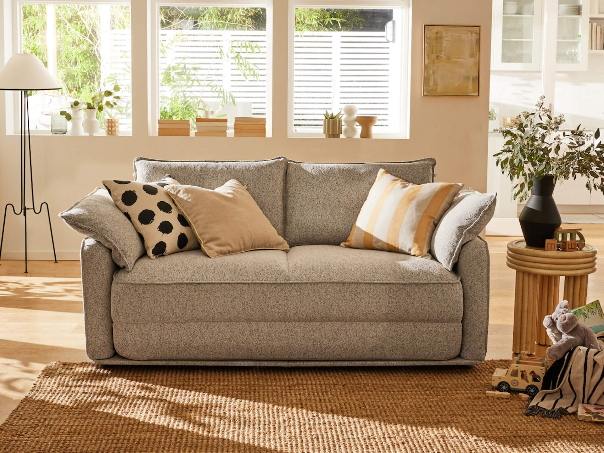 Stylish light grey sofa with assorted cushions in a sunlit living room; natural decor includes plants, wooden side tables, and a woven rug.