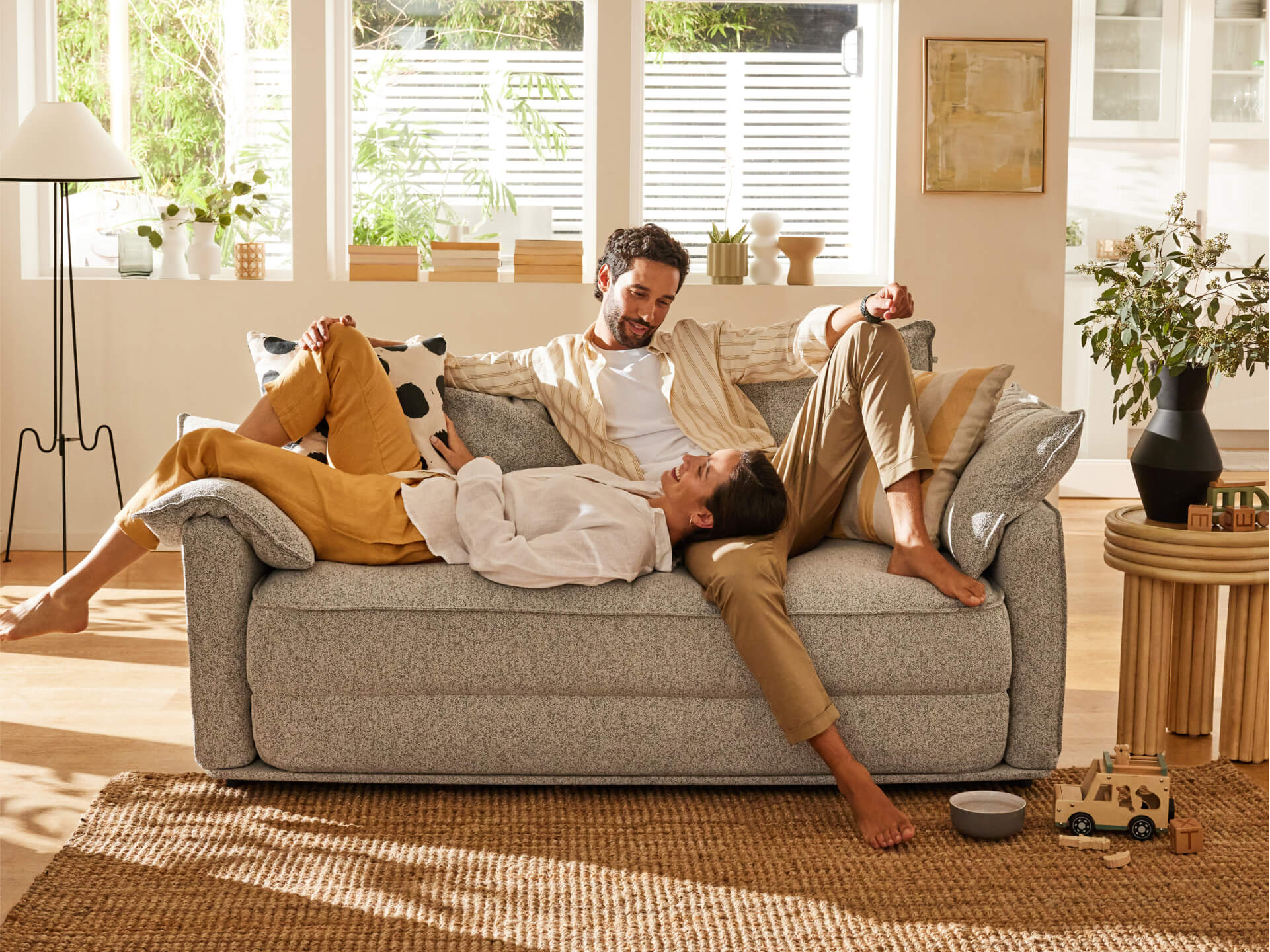 Couple lounging on a light grey sofa in a sunlit room with wooden floors, decorative plants, and modern furniture accents.