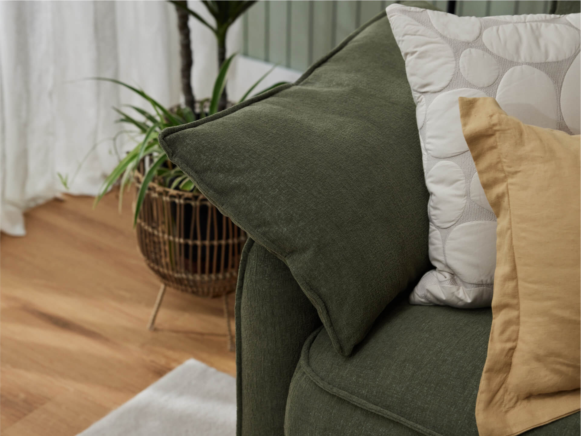 Green fabric couch with beige and patterned cushions. Wooden floor, plant in wicker basket, and white curtain in background. Cozy living room.