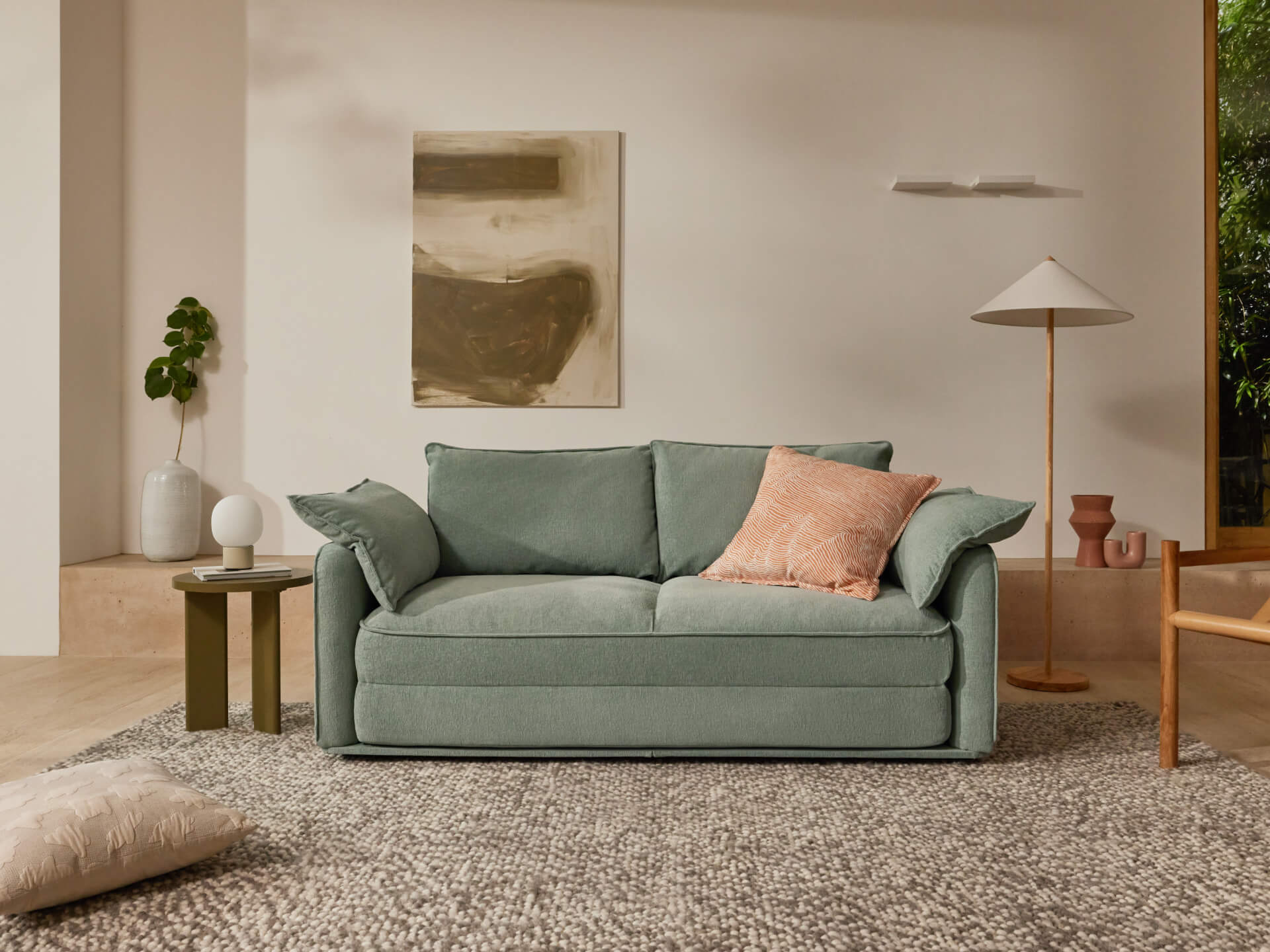 A light green sofa with cushions sits on a textured grey rug in a bright room. Nearby are a small table, lamp, and abstract wall art.
