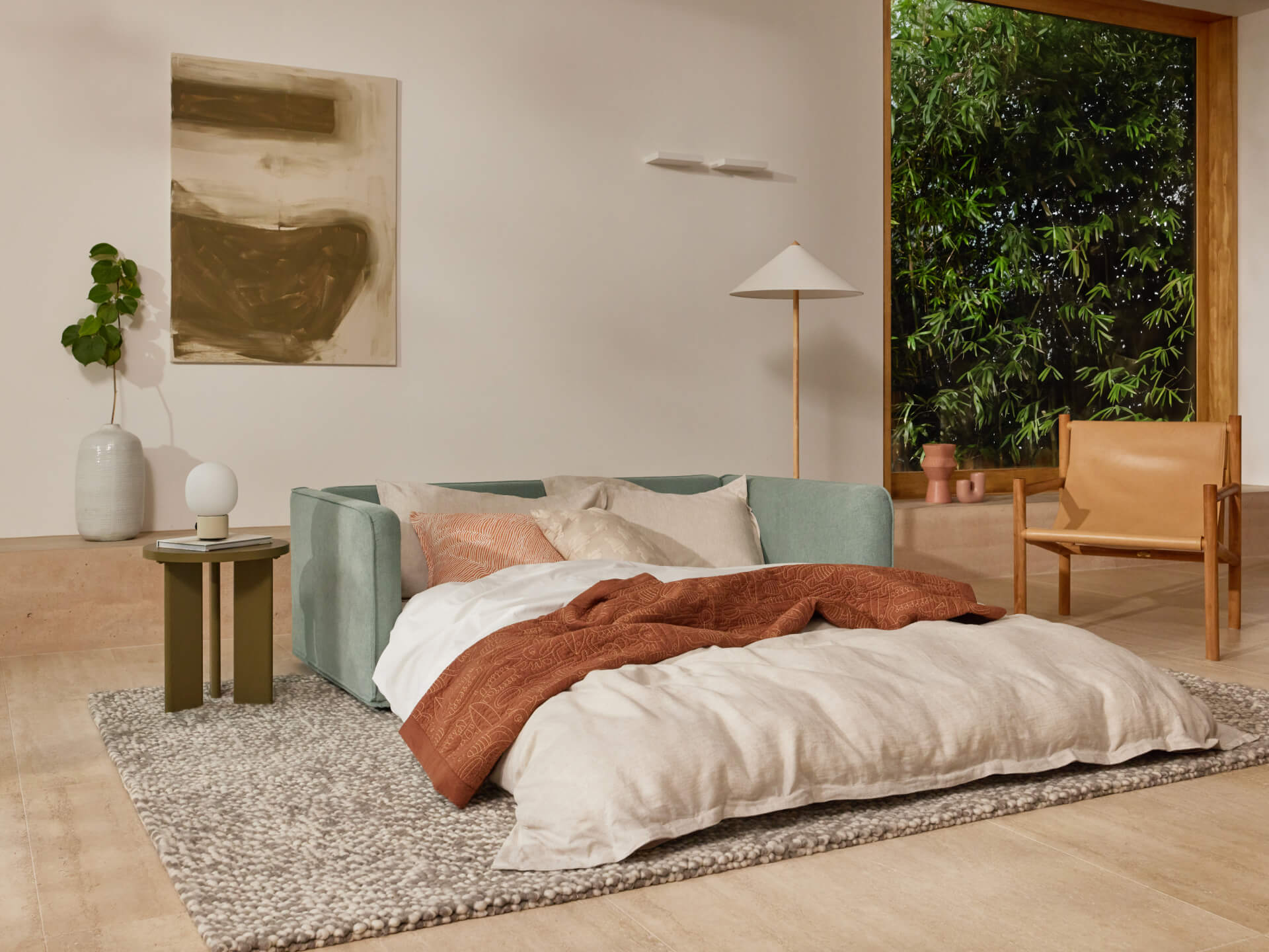Modern living room with a light green sofa bed, beige and rust bedding, wooden chair, grey rug, small green side table, and large window showing greenery.