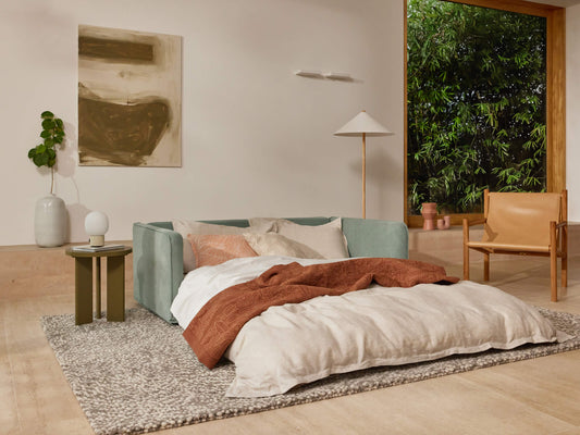 Modern living room with a light green sofa bed, beige and rust bedding, wooden chair, grey rug, small green side table, and large window showing greenery.